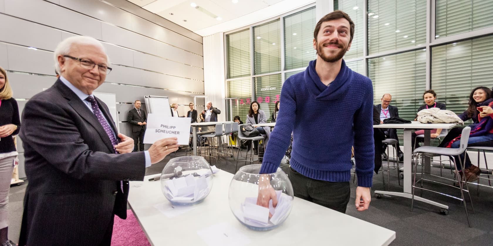 Man in a blue sweater draws a slip of paper from a glass bowl while an older man next to him holds a slip of paper with the name 'Philipp Scheucher.' Several people are seated at tables in the background.