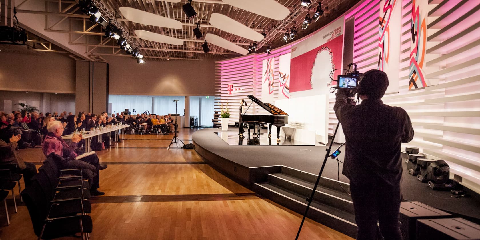 A cameraman records at an event with a piano on stage and an audience in the background.