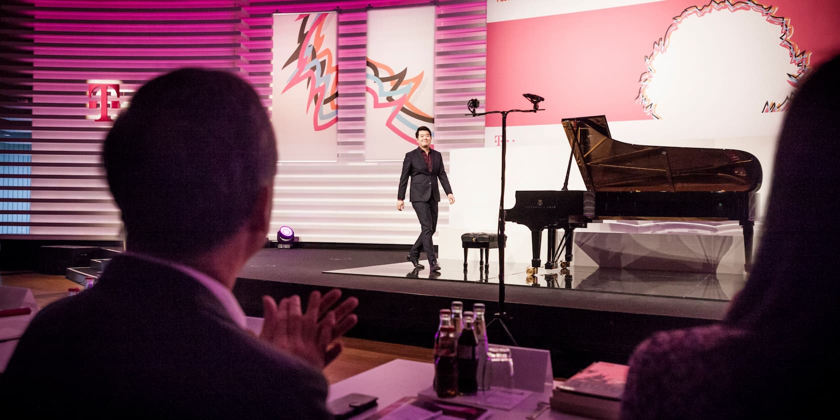 A man walks across a stage with a piano while the audience applauds. Colorful banners and the Deutsche Telekom logo are visible in the background.