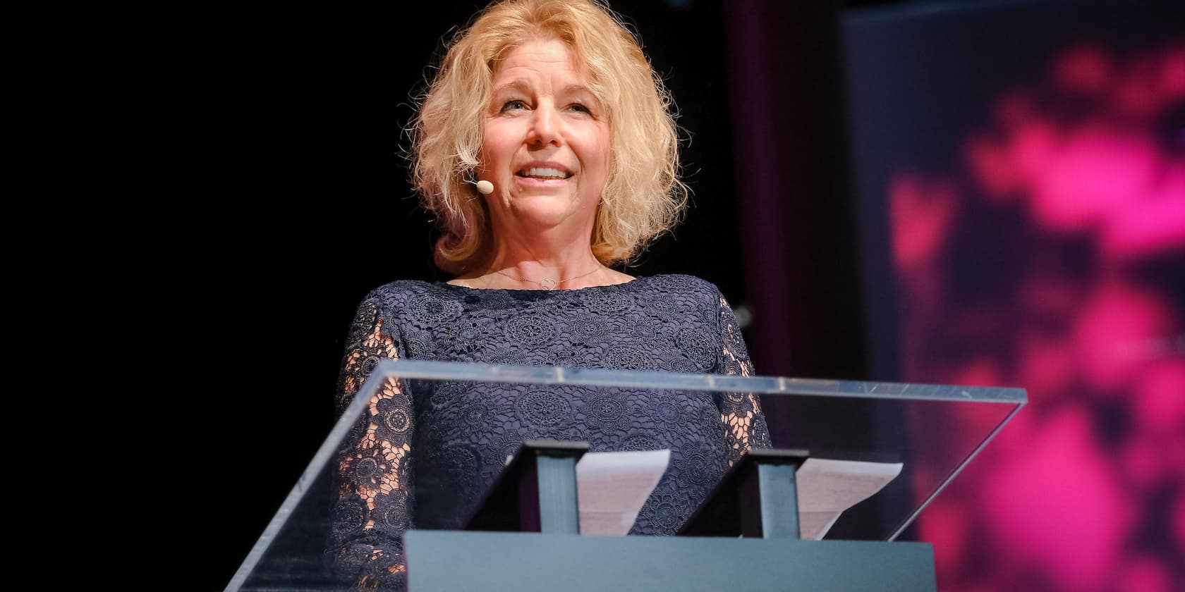 A woman with blonde hair speaks at a podium with a microphone.