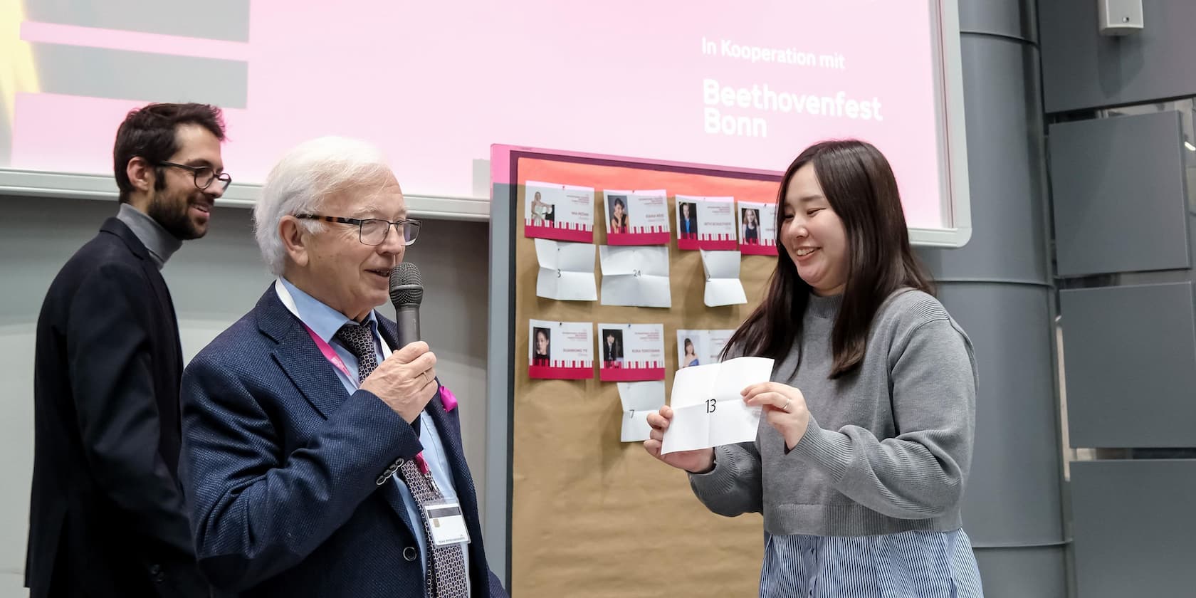 Mann mit Mikrofon überreicht einem jungen Mann ein Papier bei einer Veranstaltung mit Beethovenfest Bonn im Hintergrund.