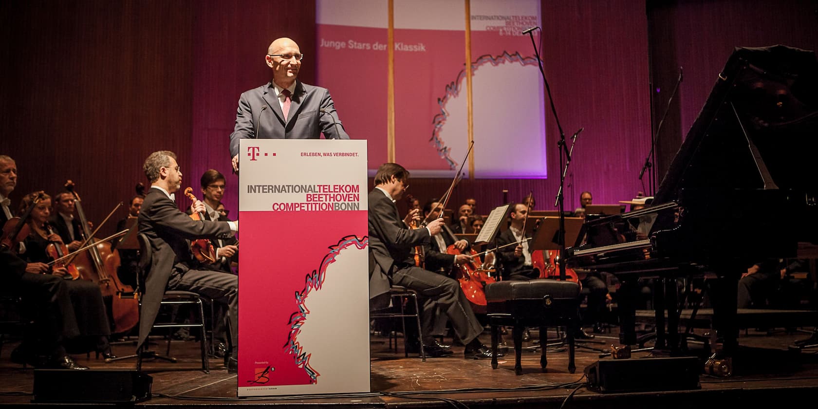 Ein Mann hält eine Rede bei der International Telekom Beethoven Competition Bonn. Im Hintergrund spielt ein Orchester.