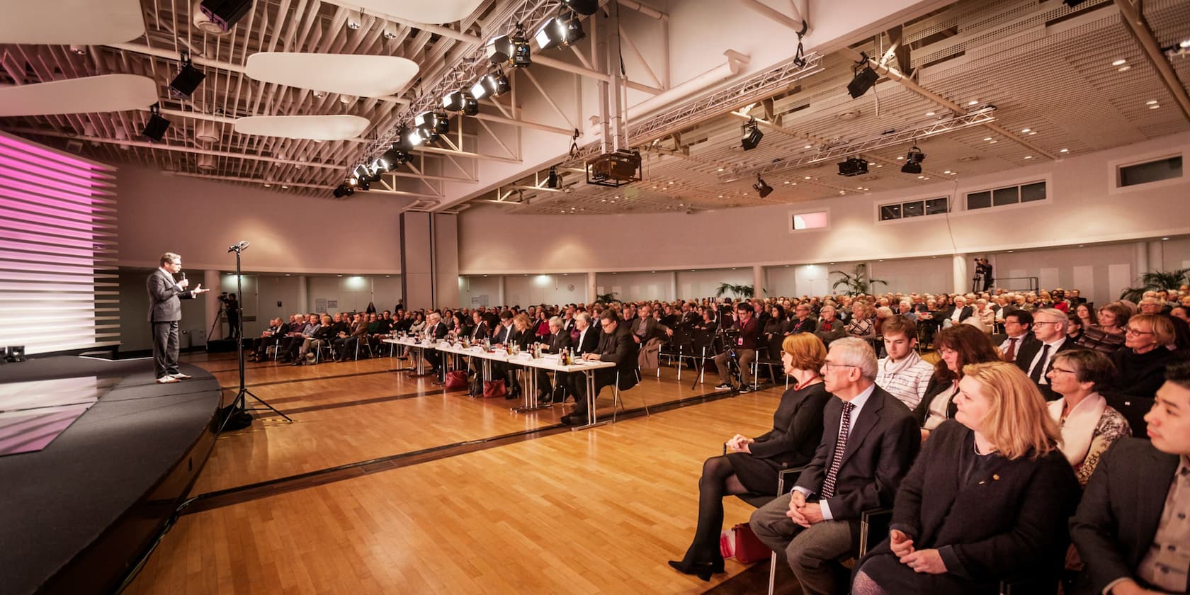 Ein Sprecher hält eine Präsentation vor einem großen Publikum in einem Konferenzsaal.