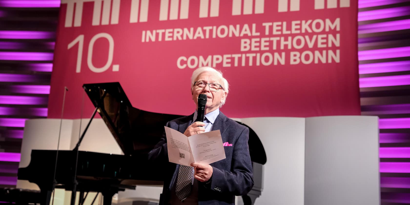 Ein Mann hält eine Rede beim 10. International Telekom Beethoven Wettbewerb in Bonn. Im Hintergrund ist ein Flügel und eine rosa Wand mit dem Veranstaltungslogo sichtbar.