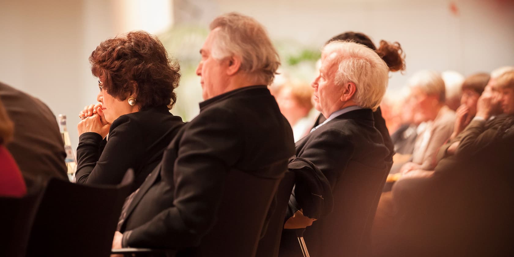 People are seated at a conference, listening attentively.