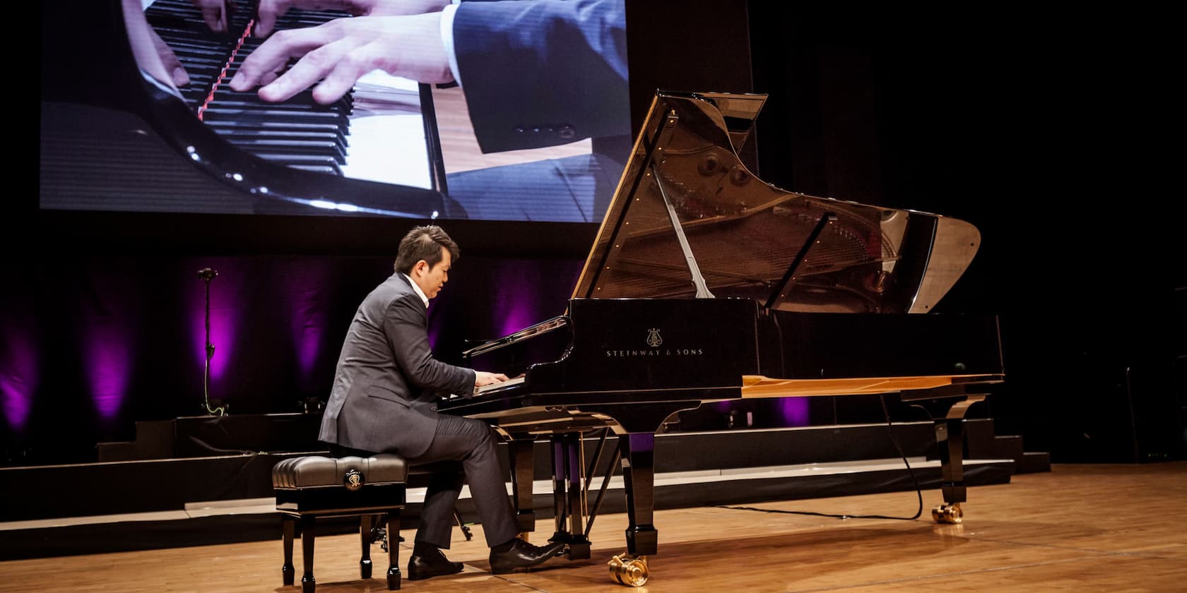 A man in a suit plays the piano on stage. In the background, a close-up of his hands on the piano is displayed on a large screen.