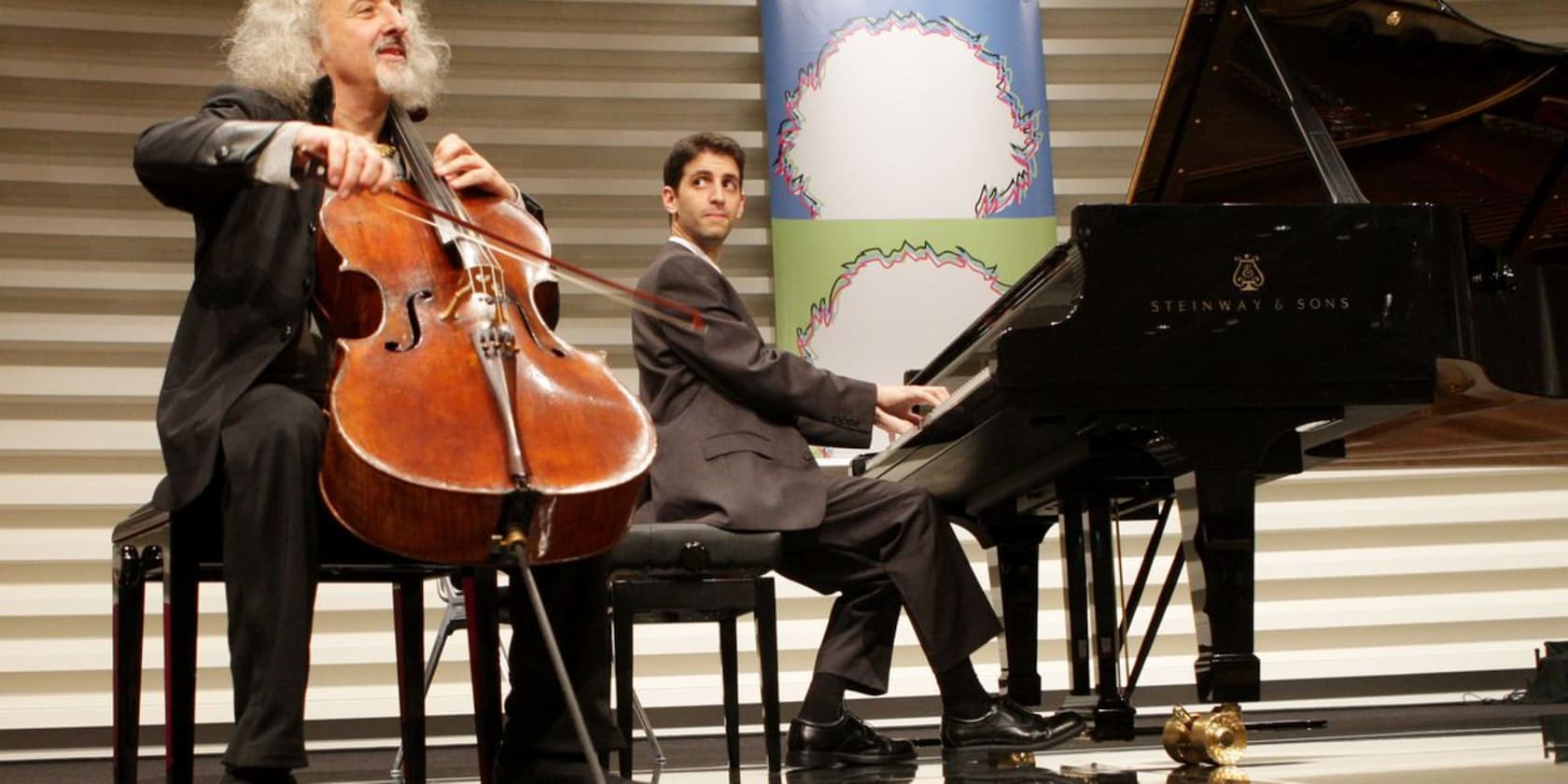 Two musicians performing on stage. One is playing the cello, the other the piano.