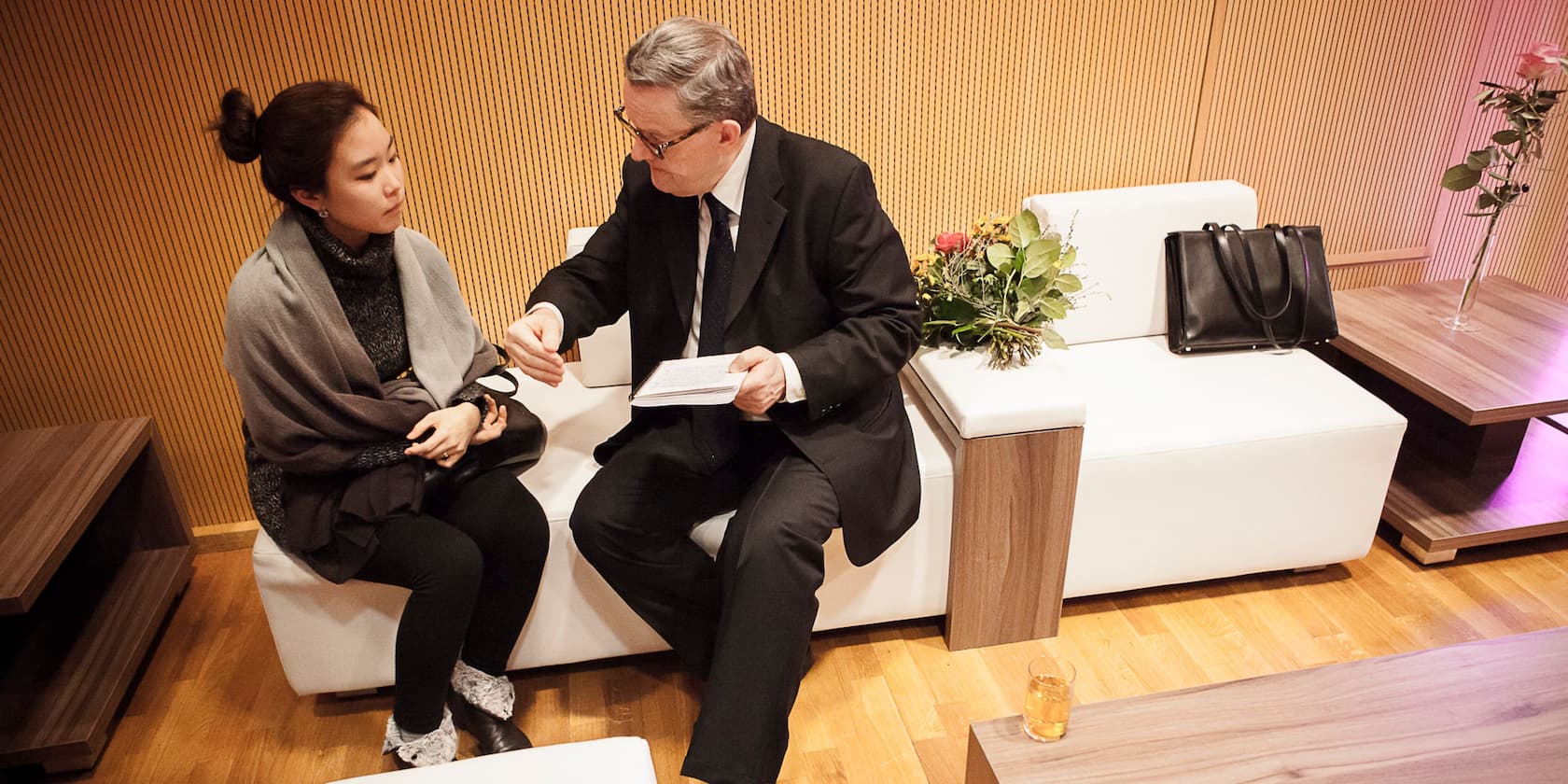 An older man and a younger woman are sitting on a white sofa, having a conversation. The man is holding a book.