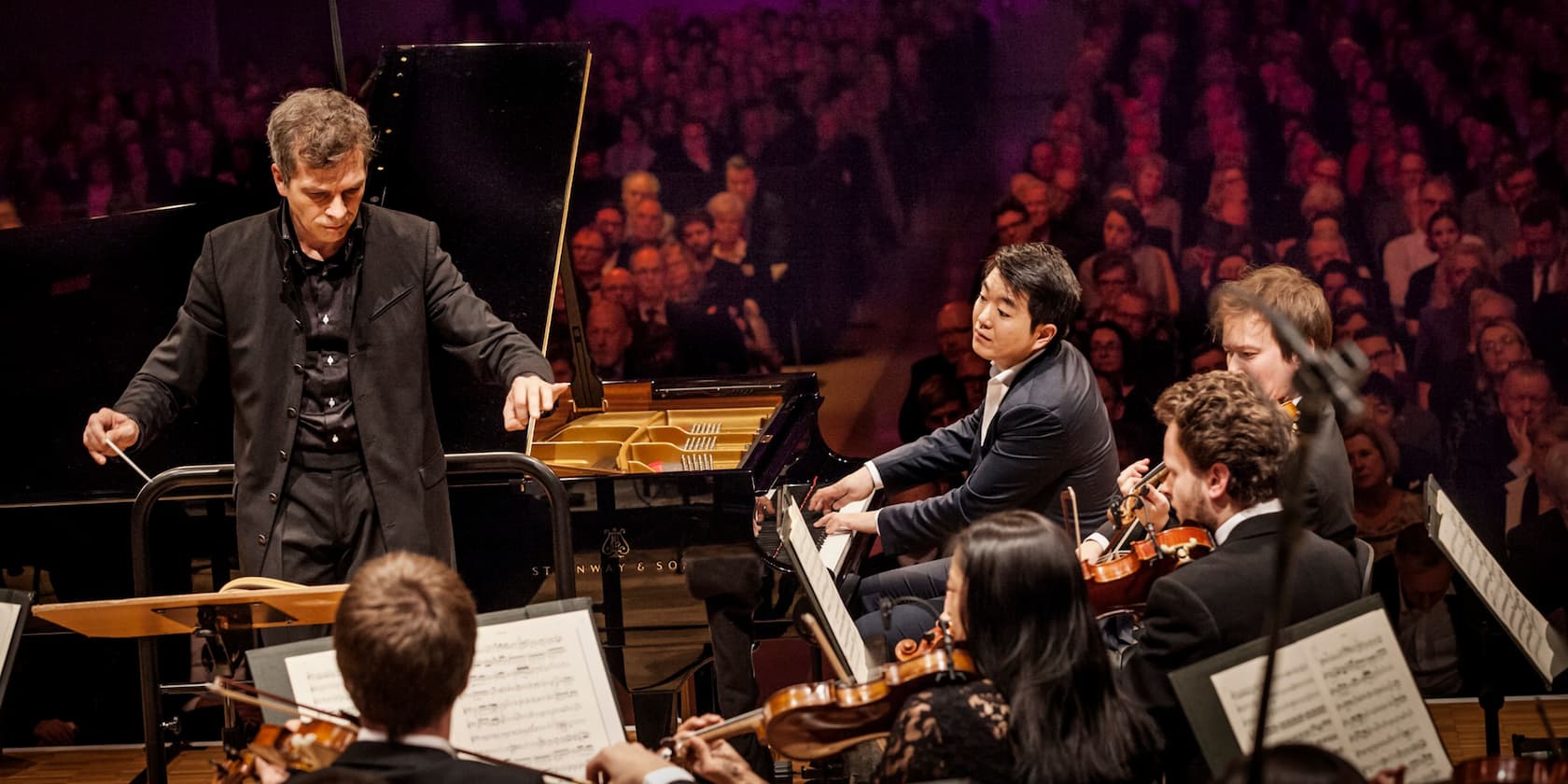 Ein Dirigent leitet ein Orchester bei einem Konzert; ein Pianist spielt im Hintergrund.