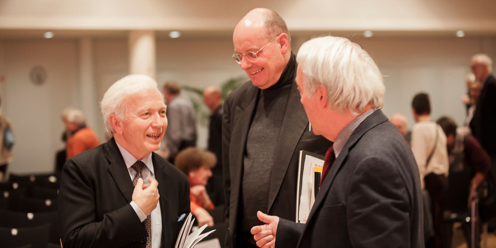 Three elderly men in suits smiling and conversing at an event.