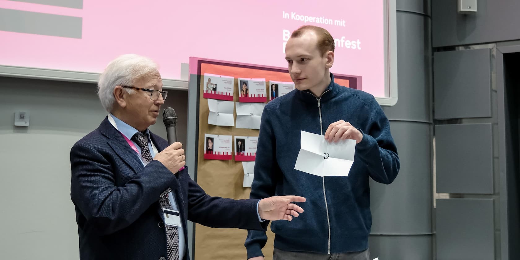 An older man with a microphone hands a piece of paper with the number 12 to a younger man. In the background, there is a board with photos and numbers.