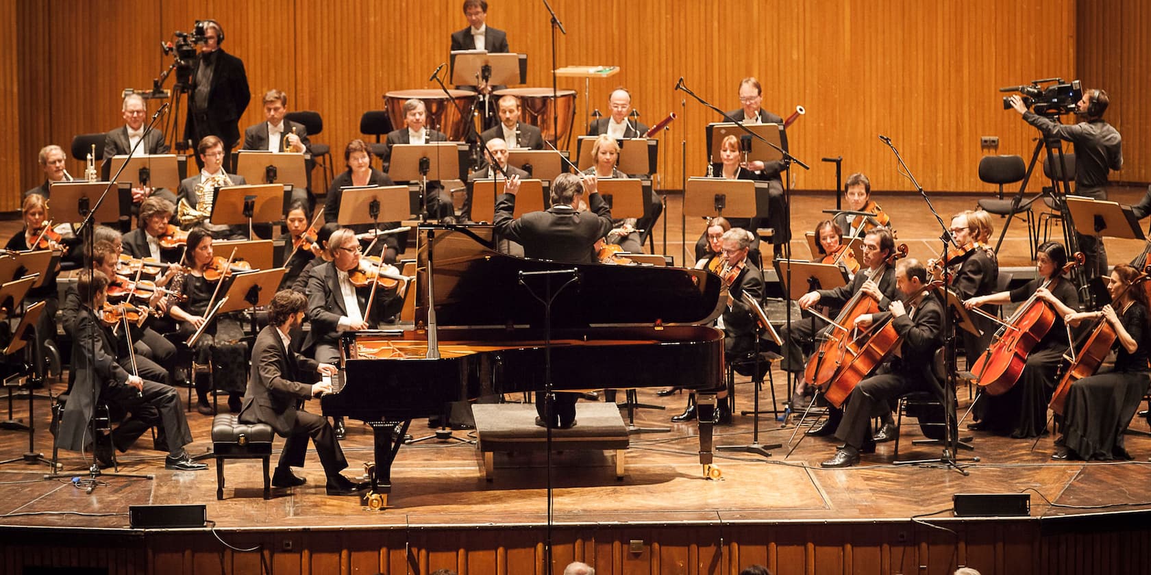 Ein Orchester und ein Dirigent führen ein Konzert auf. Im Vordergrund spielt ein Pianist am Klavier.