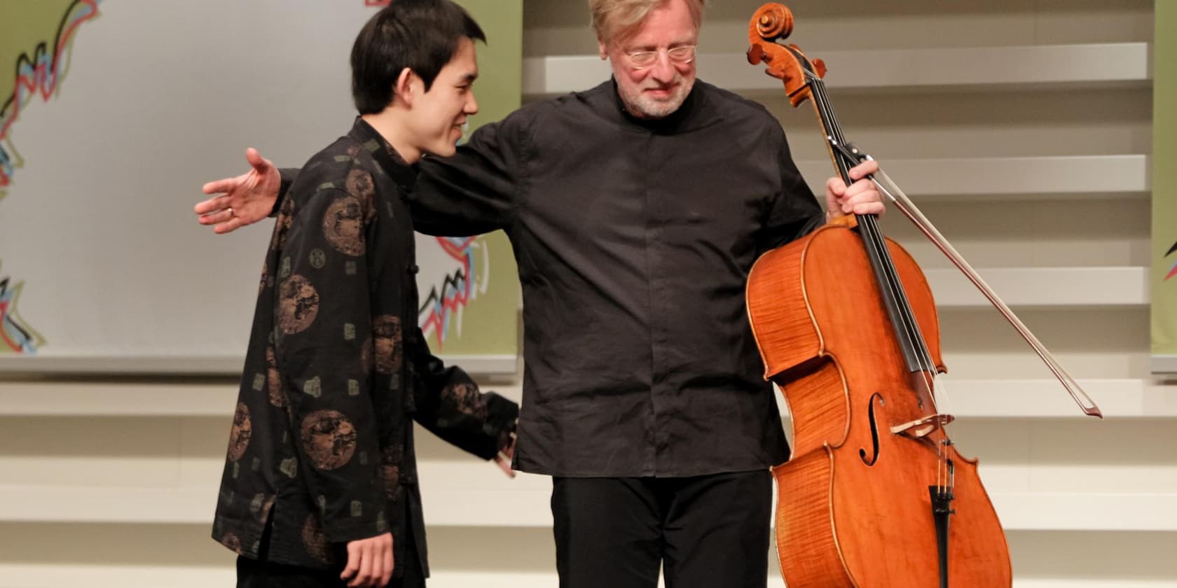 Two men on a stage, one holding a cello.