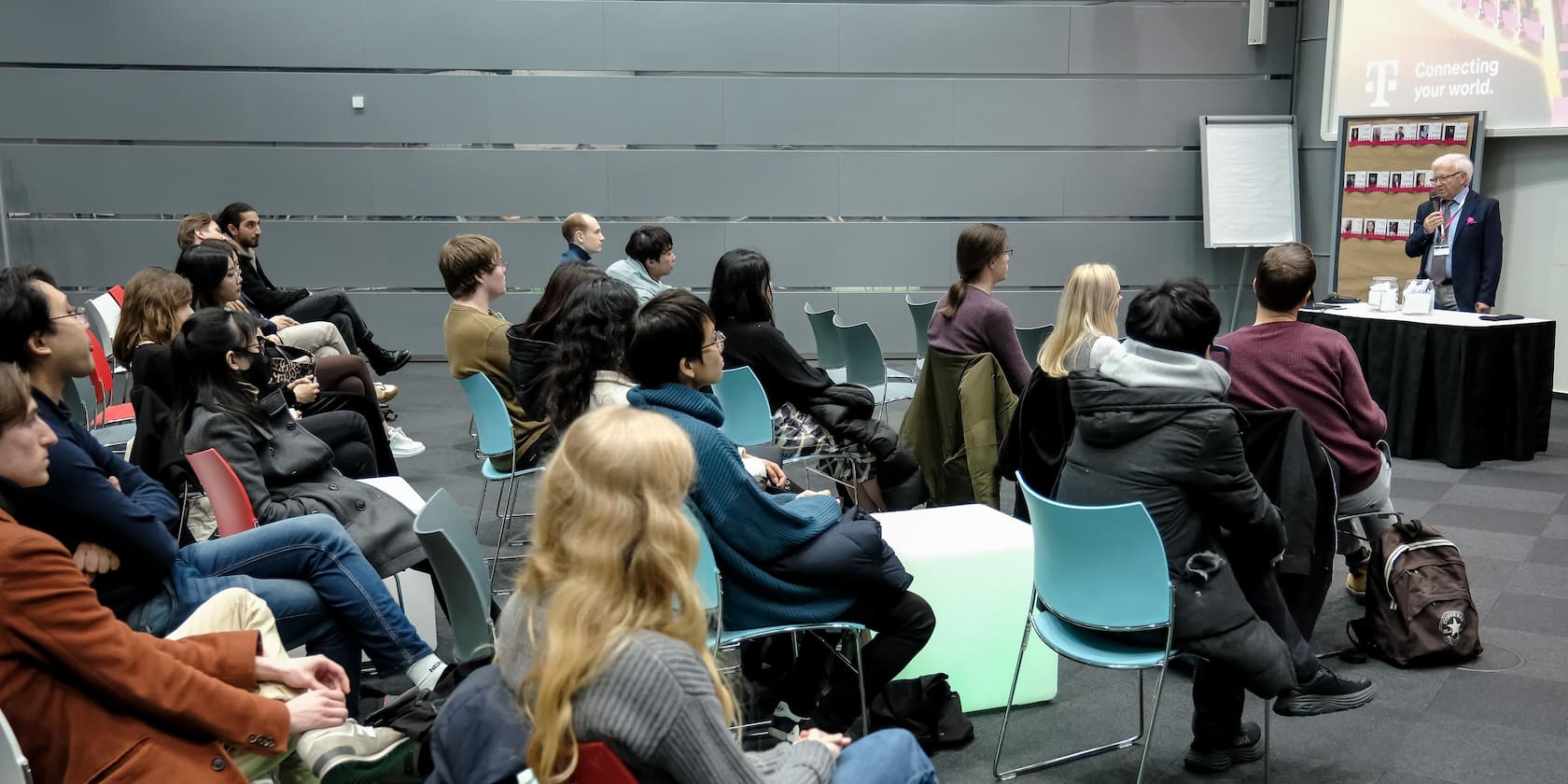 Ein Vortragender spricht vor einer Gruppe von Zuhörern in einem modernen Konferenzraum. Auf dem Bildschirm steht 'Connecting your world'.