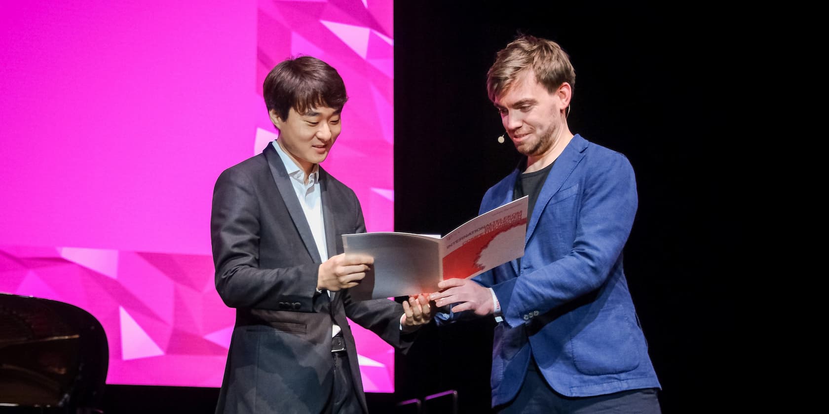 Two men in suits smile and look at a large book together on a stage with a pink background.