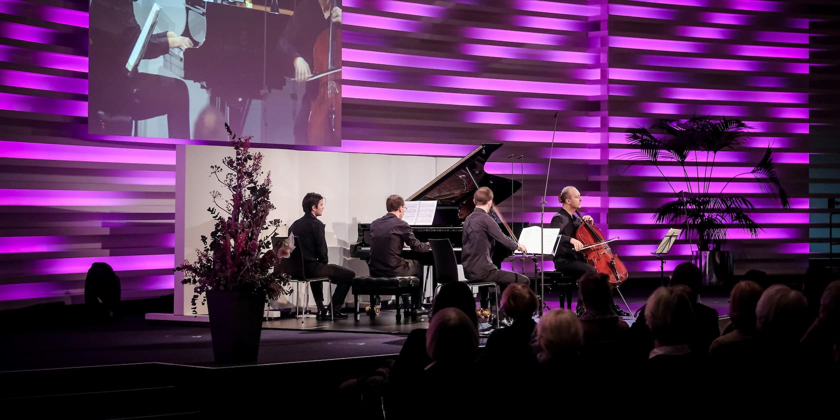 A music group performs on a stage with purple backlighting, including piano and cello.