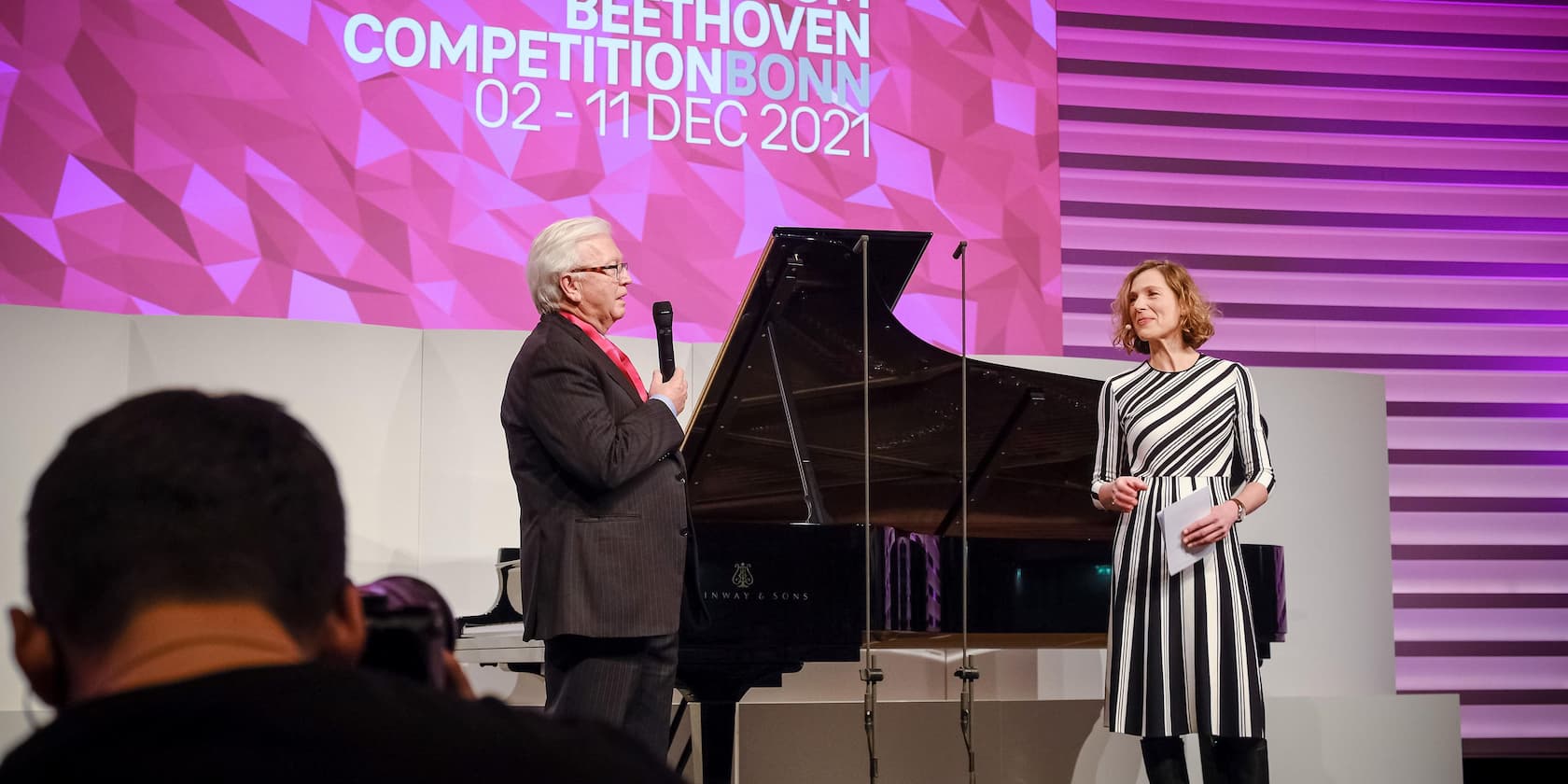 A man and a woman stand next to a piano and speak during the Beethoven Competition Bonn 2021.