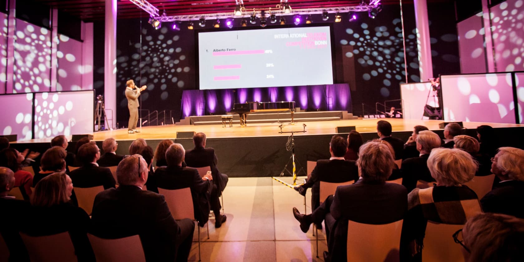 People in an auditorium watch a brightly lit stage with a large screen presentation.