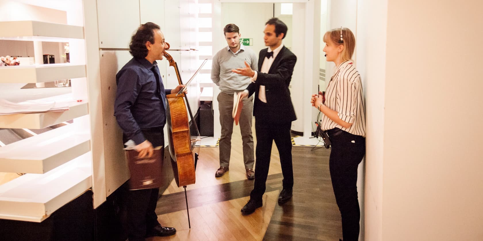 Four people are talking in a hallway. One person is holding a cello.