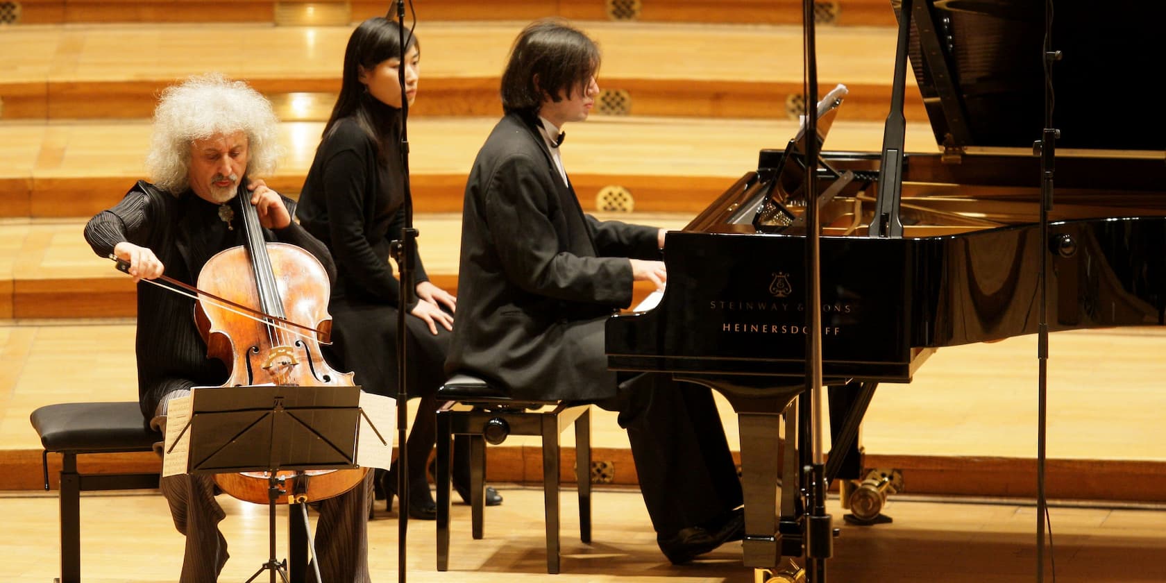 A cellist performs in front of a grand piano being played by a pianist in a concert hall.