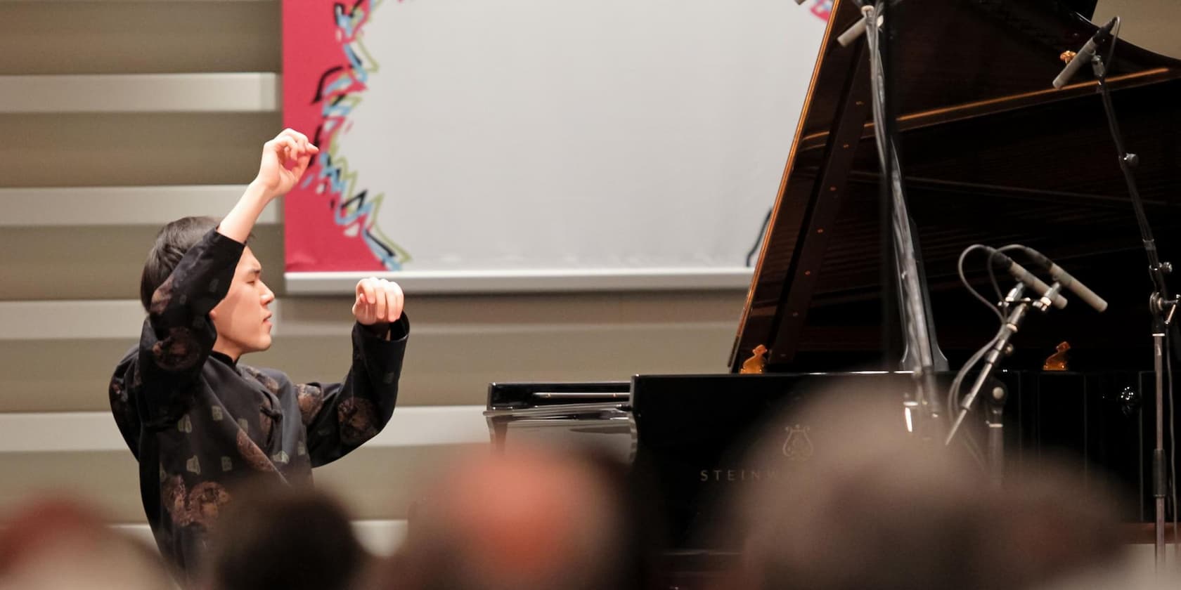 A musician thoughtfully conducts beside a grand piano on stage.