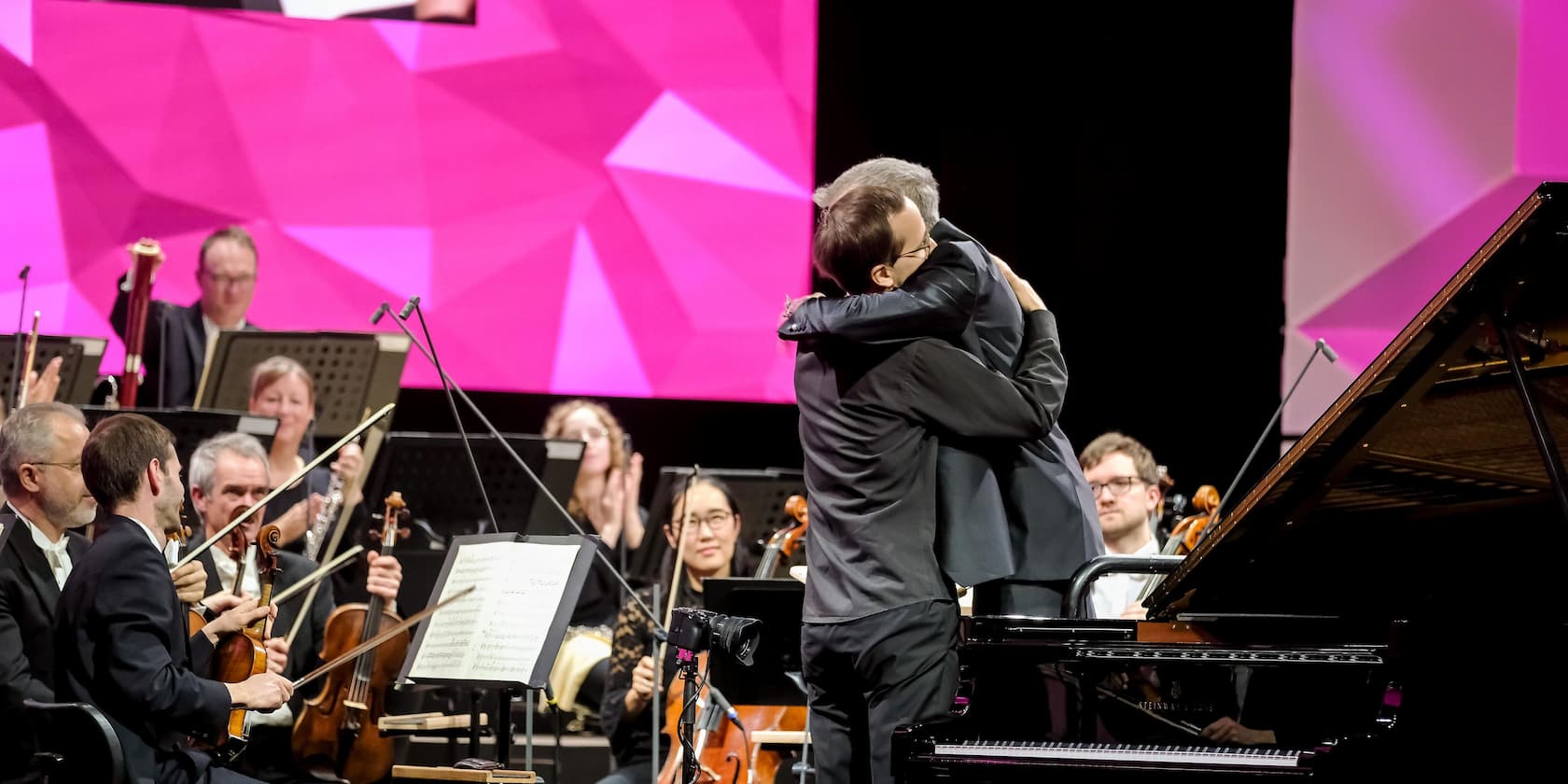 Conductor and pianist embrace in front of an orchestra on stage.