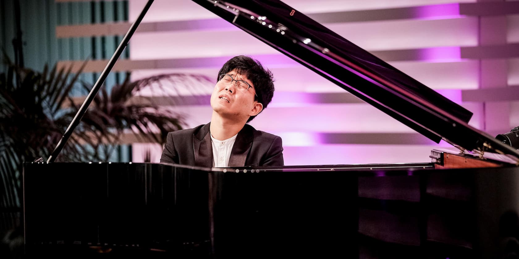 Musician playing a grand piano during a concert, with closed eyes and purple background lighting.