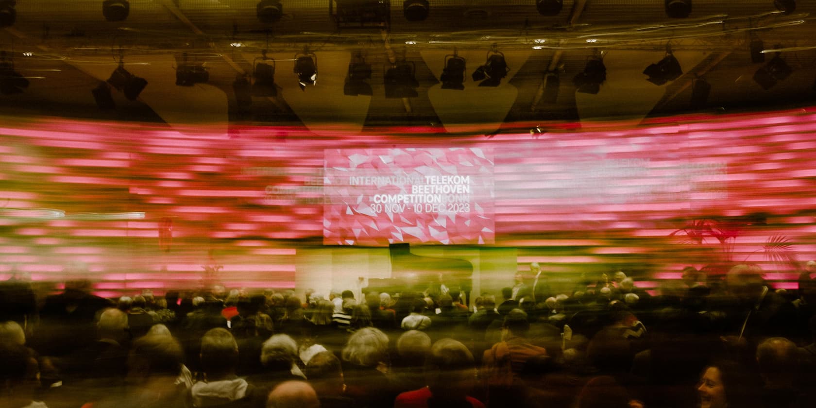 Blurred image of audience at the International Telekom Beethoven Competition Bonn with illuminated stage backdrop. Text on backdrop: International Telekom Beethoven Competition Bonn, 30 Nov - 10 Dec 2023.