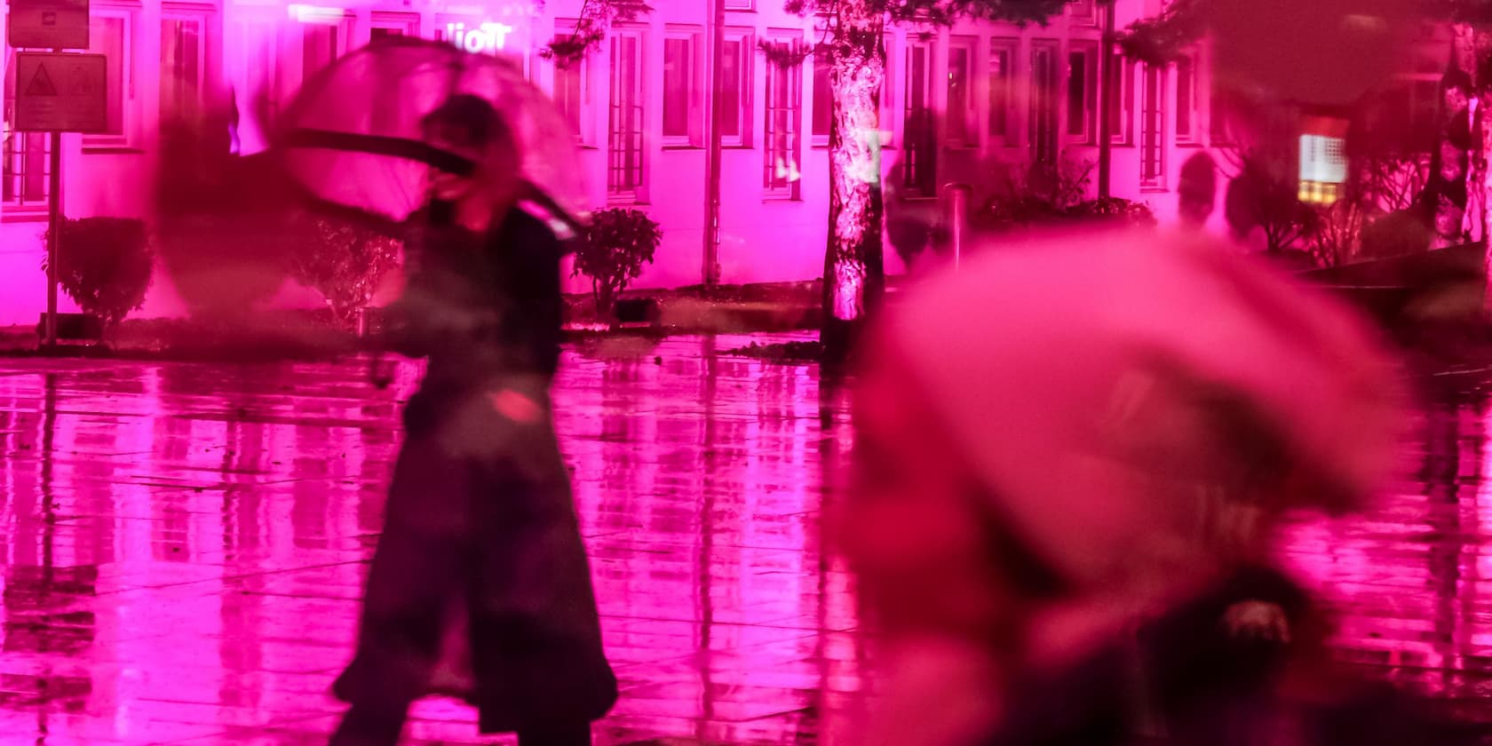 People with an umbrella and a hat walk through a street illuminated in pink light in the rain.