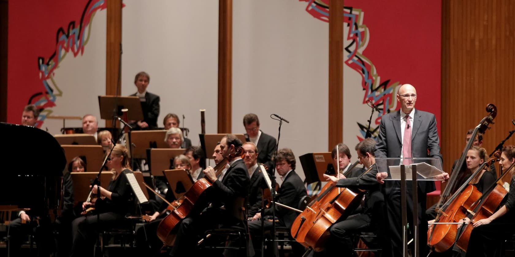 Ein Mann im Anzug hält eine Rede vor einem Orchester in einem Konzertsaal.