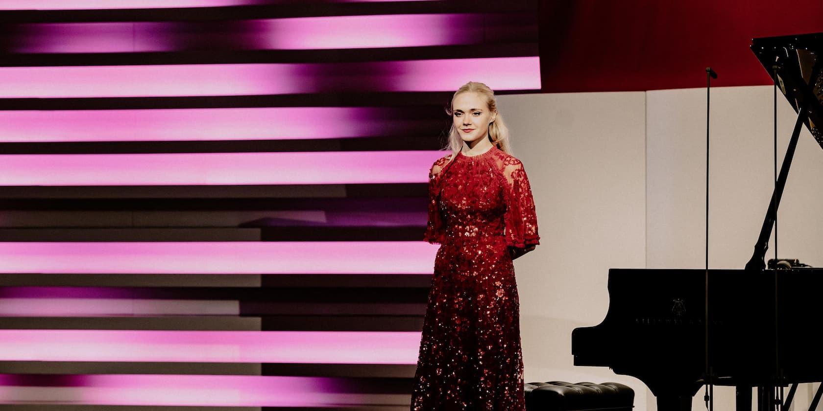 A woman in a red dress stands next to a grand piano, in front of a background with pink and white stripes.