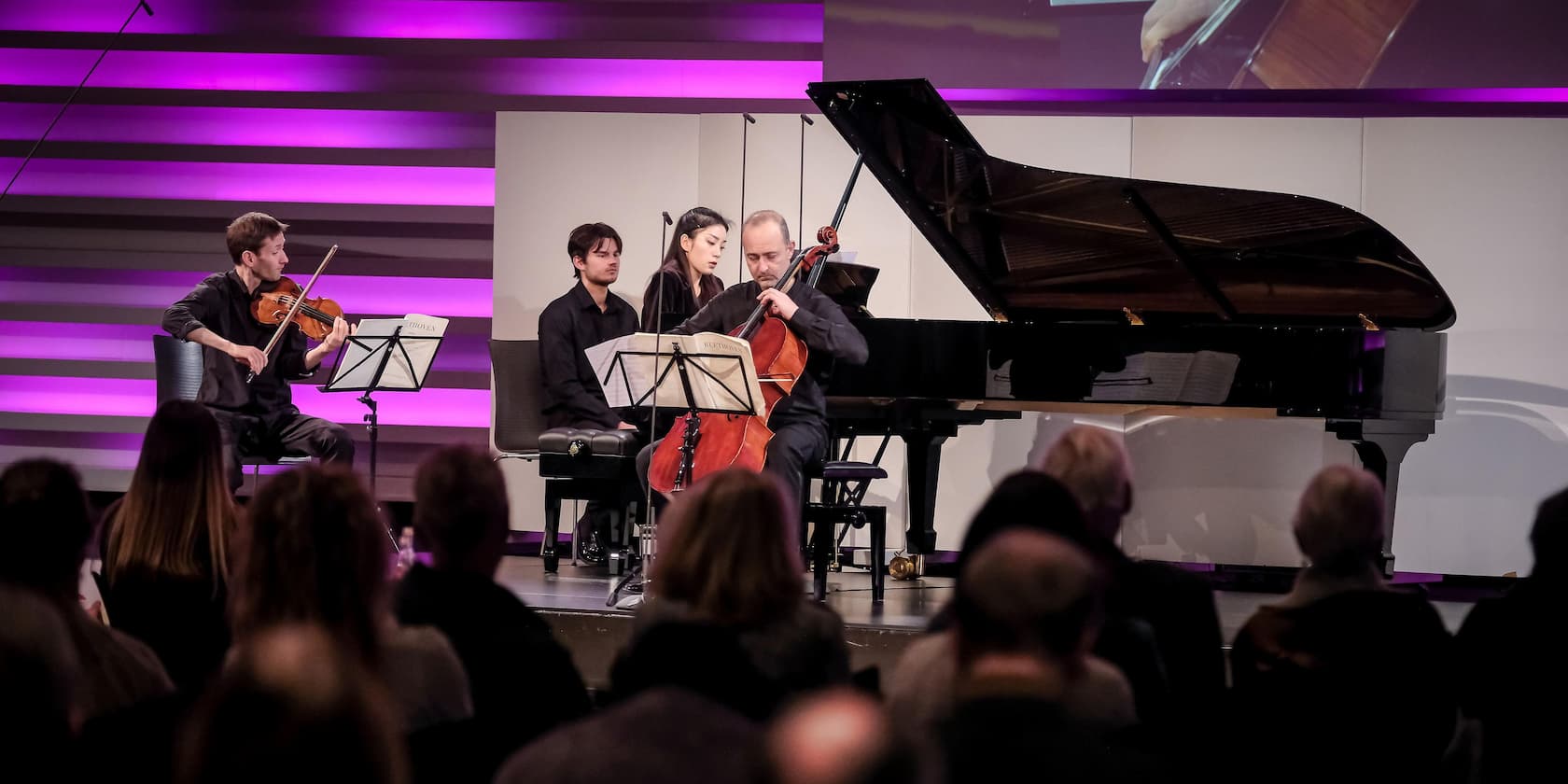 A string quartet performs on stage in front of an audience, with purple-pink background lighting.