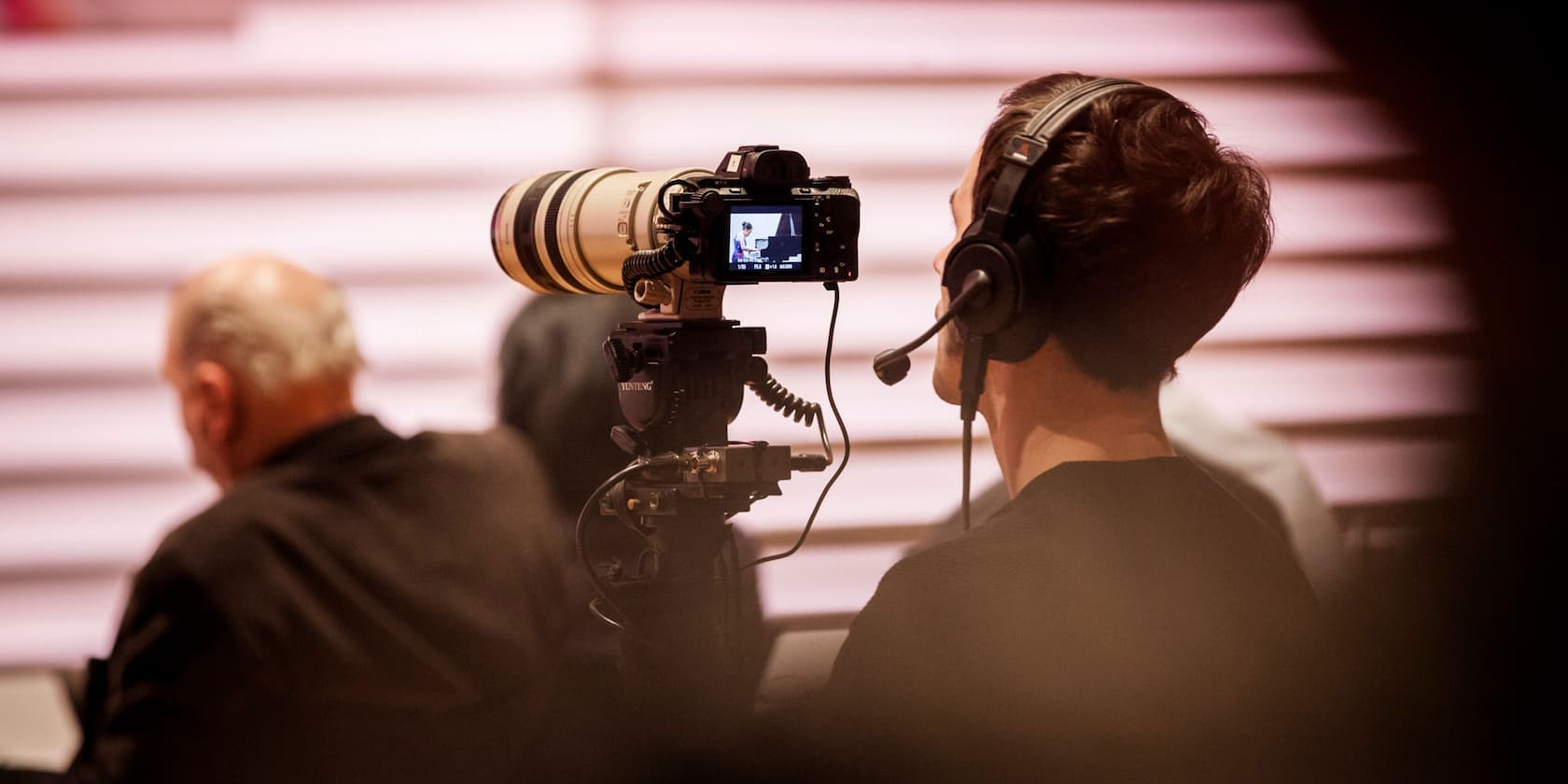 A cameraman is filming an event. A person is seated in the foreground.