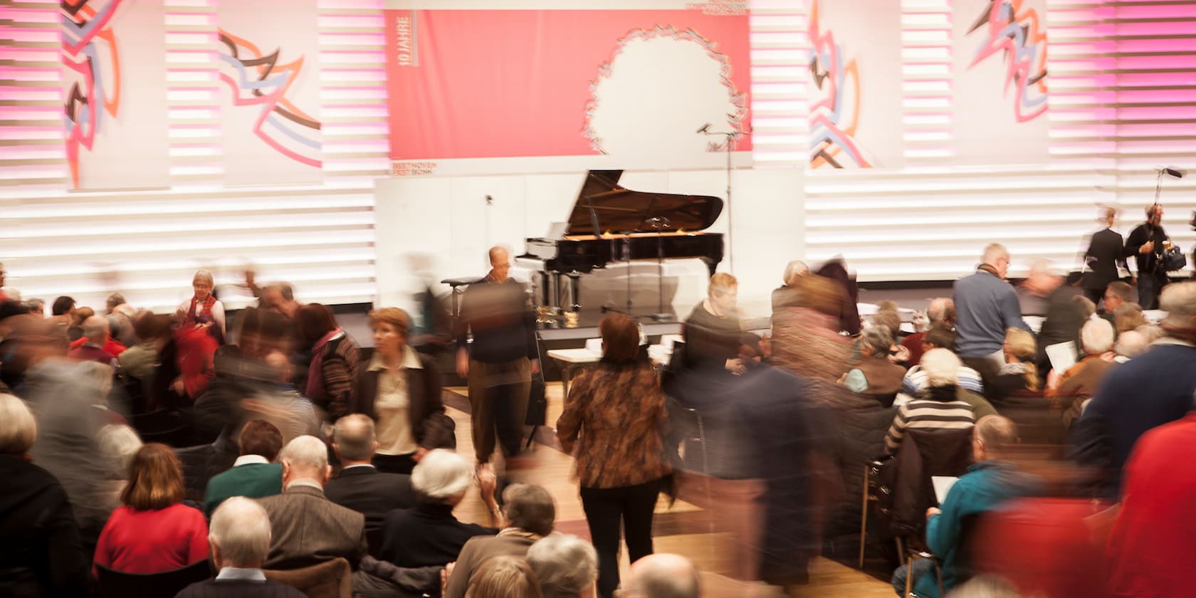 View of an audience in an event hall with a grand piano in the background.