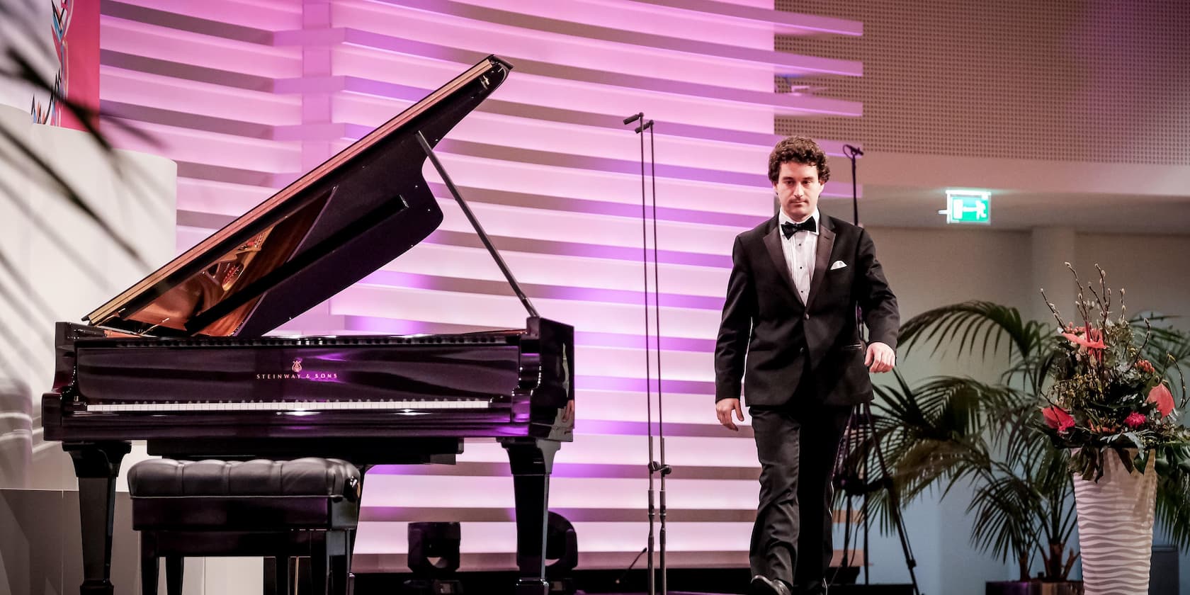 A man in a tuxedo walks past a grand piano on a stage with purple lighting.