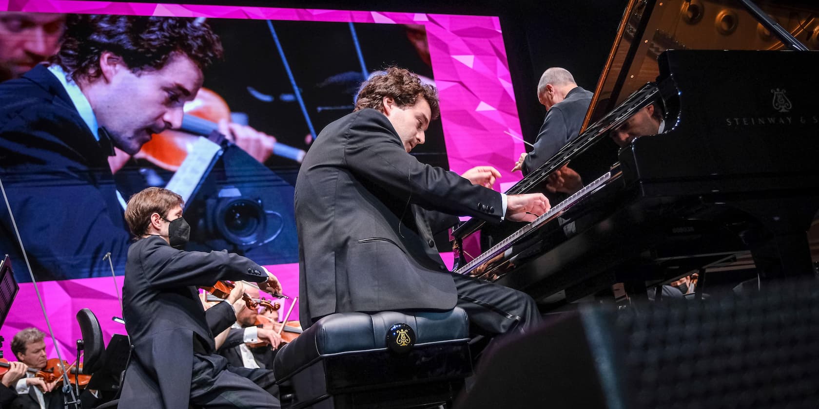 A pianist is playing on a Steinway grand piano while a violinist is visible in the foreground. A close-up of the pianist is displayed on a screen in the background.