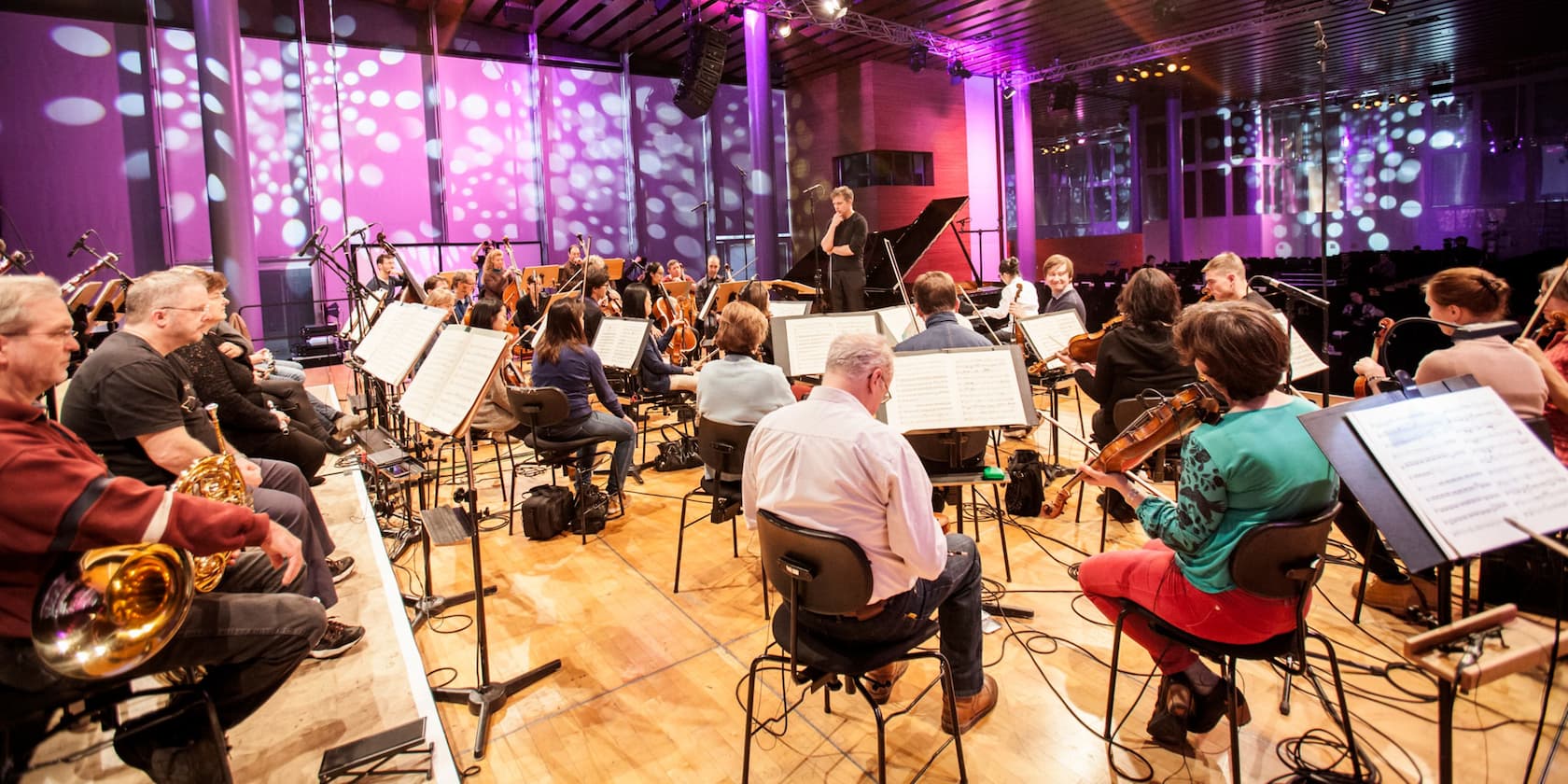 An orchestra rehearses on a stage with bright lights in the background.