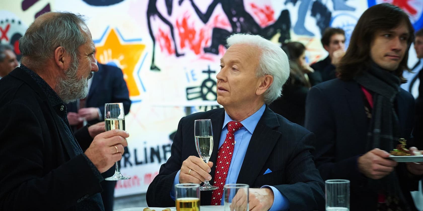 Two older men chatting over a glass of champagne, with people in the background in front of a painted wall.