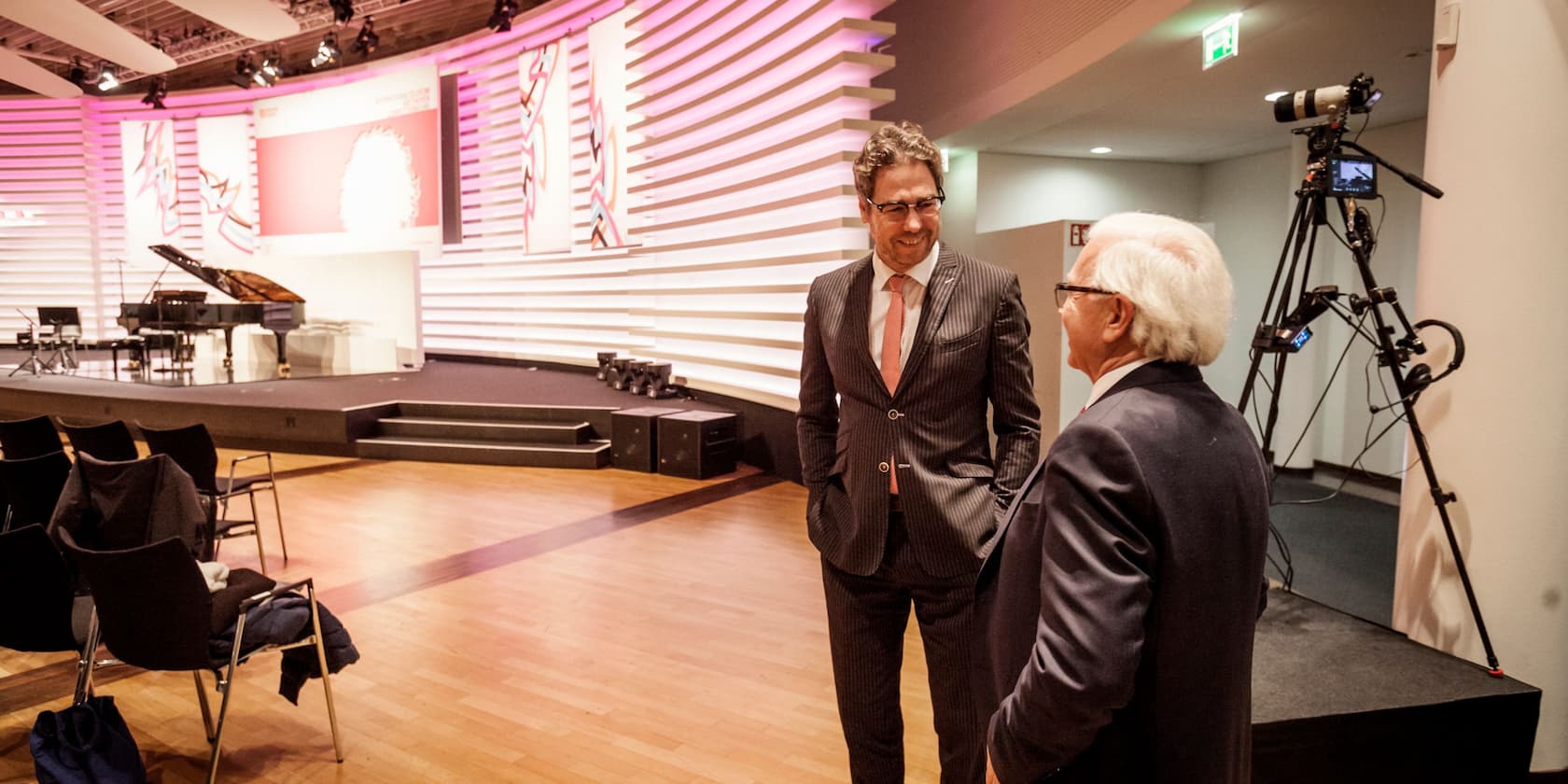 Two men in suits are conversing on a stage in front of a piano and chairs. A camera on a tripod is in the background.