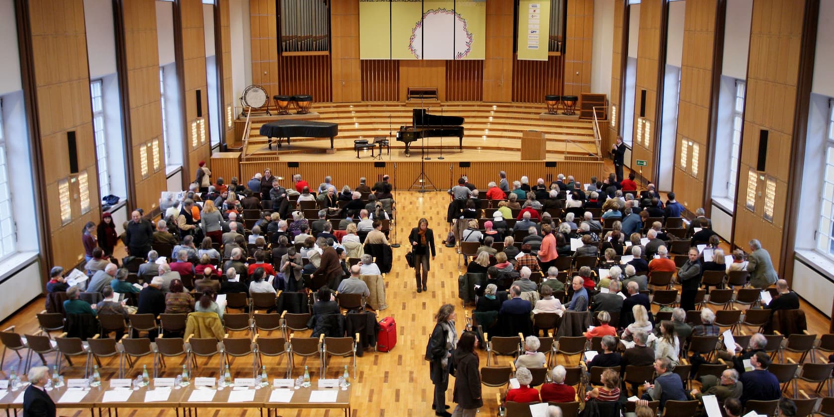 People gathering in an auditorium with a stage, two pianos, and music notation displays.