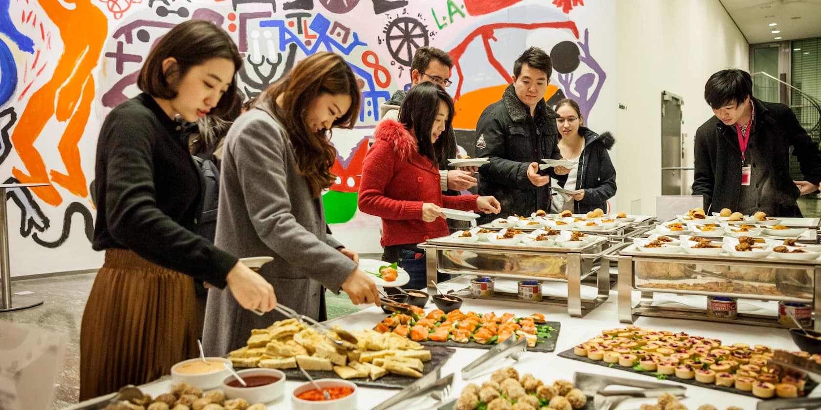 Menschen stehen an einem Buffet mit verschiedenen Vorspeisen, vor einer Wand mit bunten Kunstwerken.