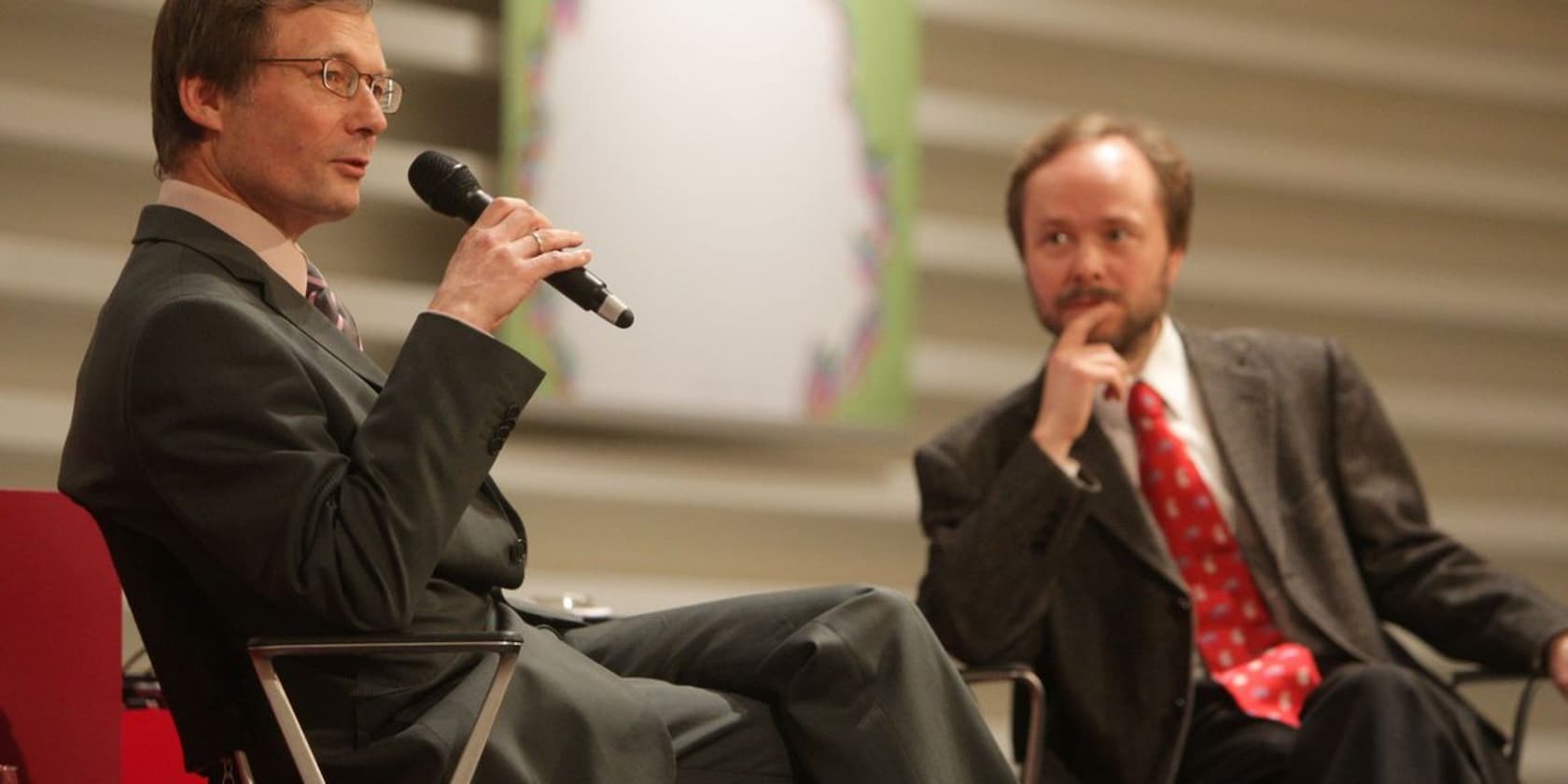 Two men in suits, one speaking into a microphone, the other listening.