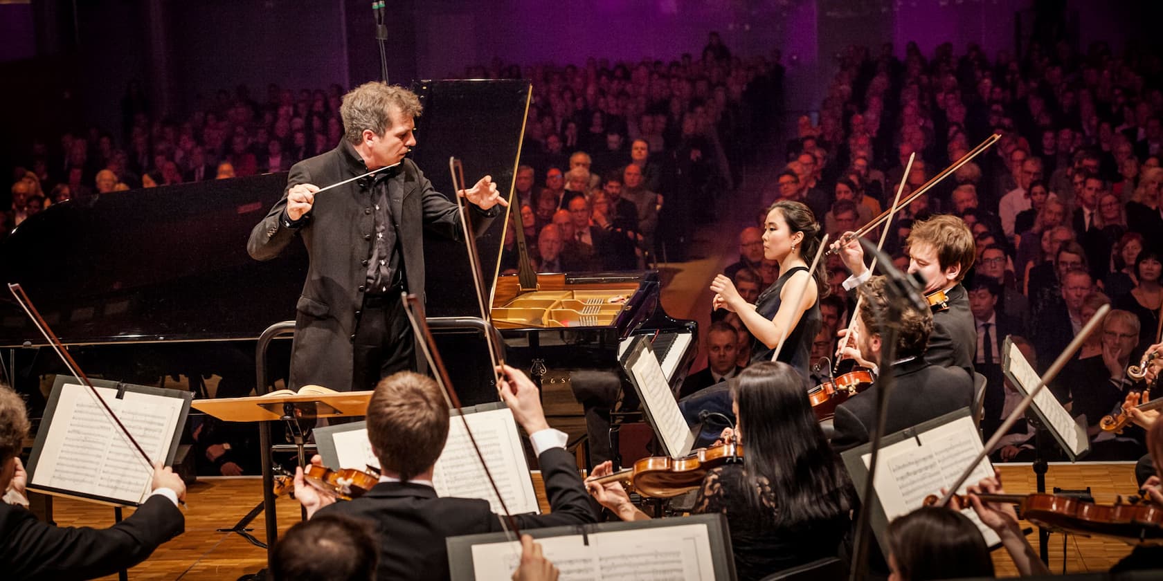 A conductor leads an orchestra while a pianist plays in the background. The audience is seated and watching.