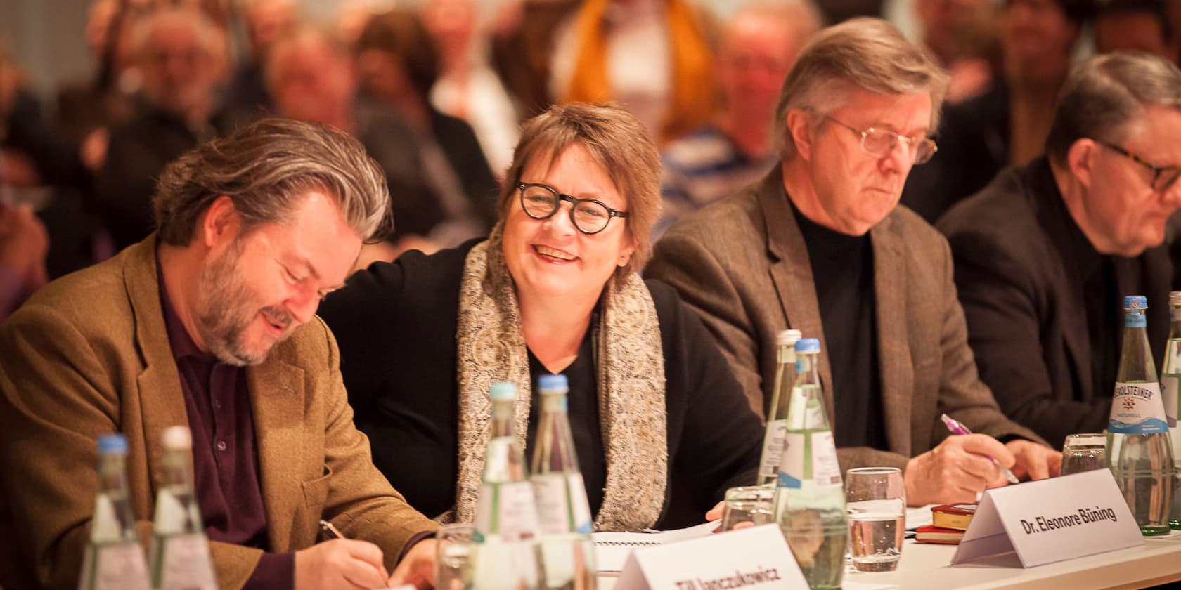 Several people are sitting at a conference table, taking notes. A woman in the center is smiling.