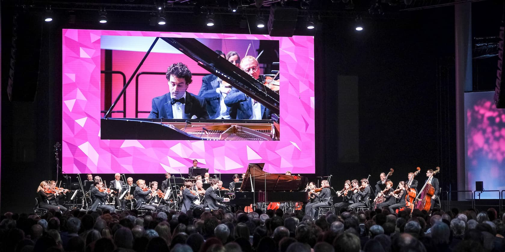 Ein Orchester spielt ein Konzert vor einem Publikum. Ein Pianist im Anzug spielt auf einem Flügel, und eine Nahaufnahme davon wird auf einer großen Leinwand im Hintergrund angezeigt.