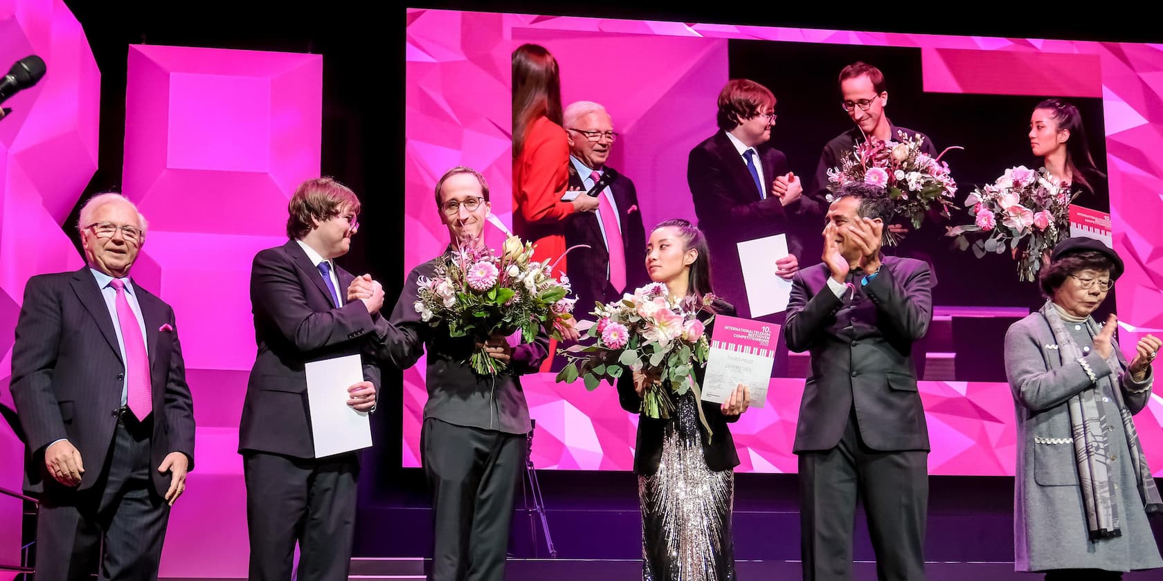 Award ceremony with several people on a stage. Two individuals are holding bouquets and certificates. The scene is shown again on a screen in the background.