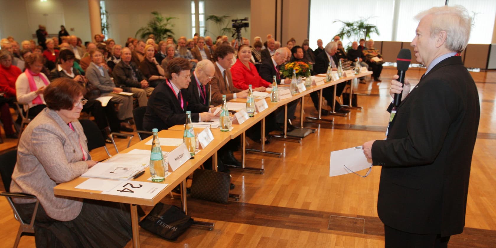 A man speaks into a microphone in front of an assembly of people. Several individuals are seated at a long table in the front, while the audience sits in the background.