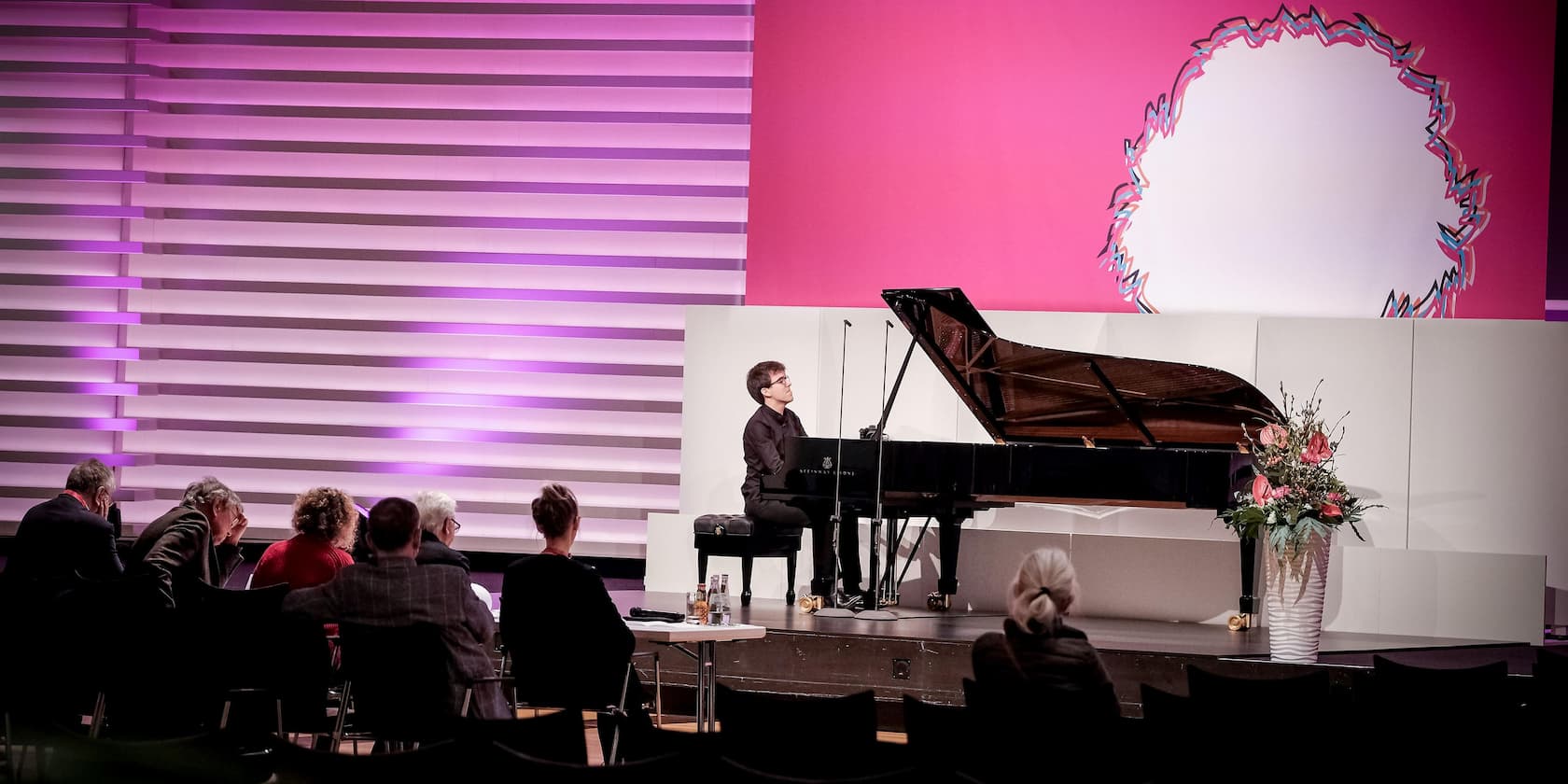 A man plays the piano on a stage in front of a small audience. The background is illuminated in pink and purple.