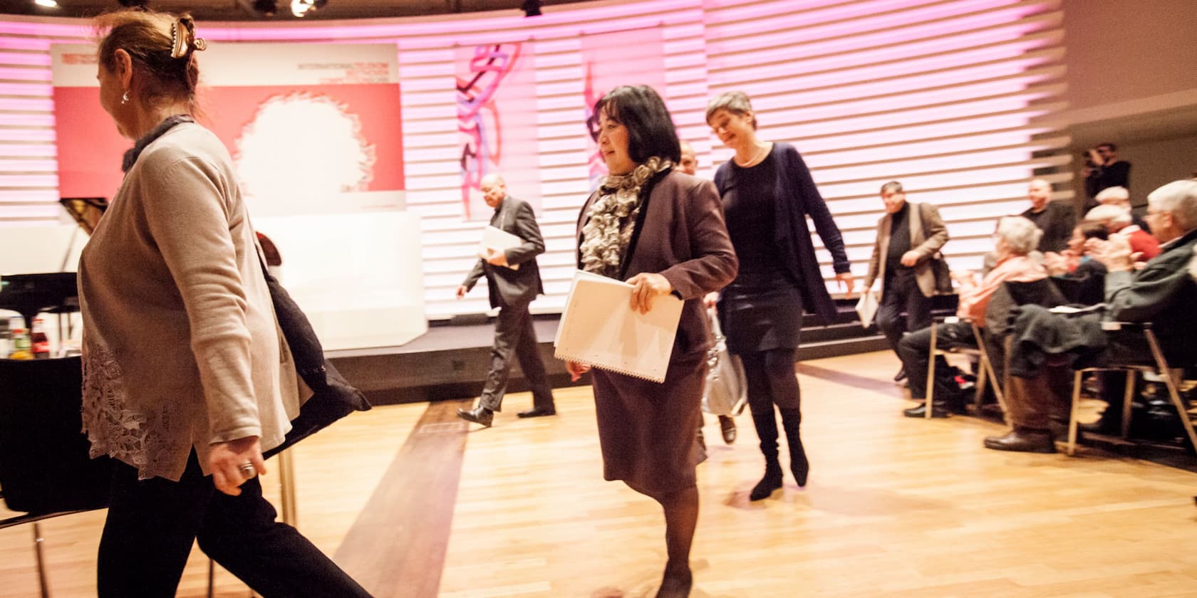 Several people walk past a seated audience in a conference room.