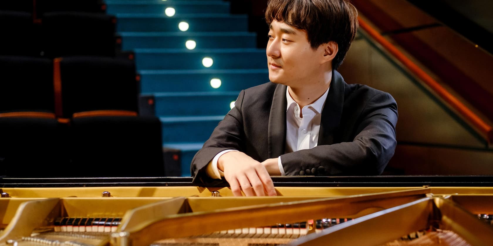 A man sits at a piano in a concert hall, looking to the side.